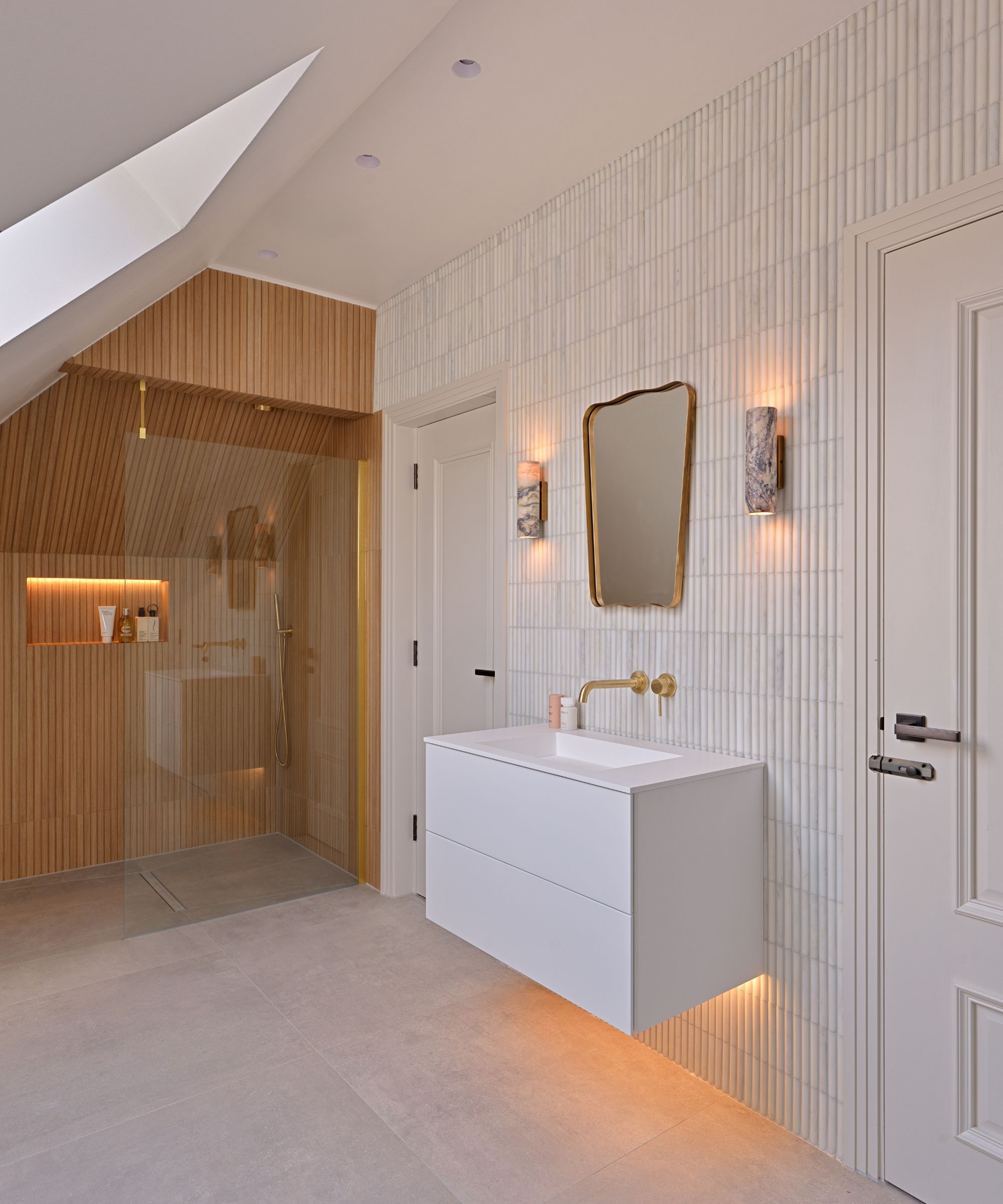 A warm, elegantly lit attic bathroom combining oak slat-clad shower walls with amber LED accent lighting and slim white vertical wall tiles. A white floating vanity with an integrated basin and brass wall tap sits below a brass-framed asymmetric mirror flanked by two stone wall sconces.