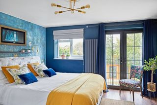 Bedroom with blue painted wall, feature wall with blue wallpaper, bed with white bedding and yellow throw, and Juliet balcony dressed with blue curtains