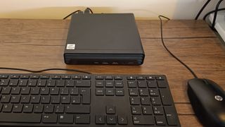 A black compact HP EliteDesk 800 desktop PC sitting on a brown wooden table in front of a black HP keyboard and a black HP mouse