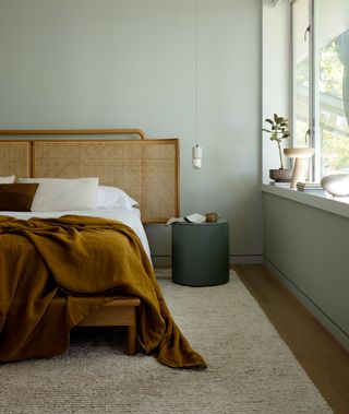 Bedroom with safe walls, brown comforter on white sheets and sage green walls with round bedside table and bright windows with pottery and plants on the ledge