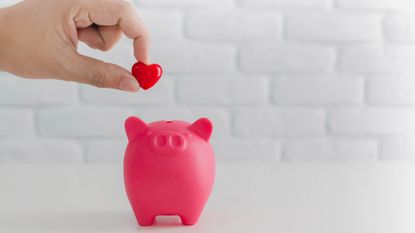 A hand deposits a heart into a pink piggy bank.