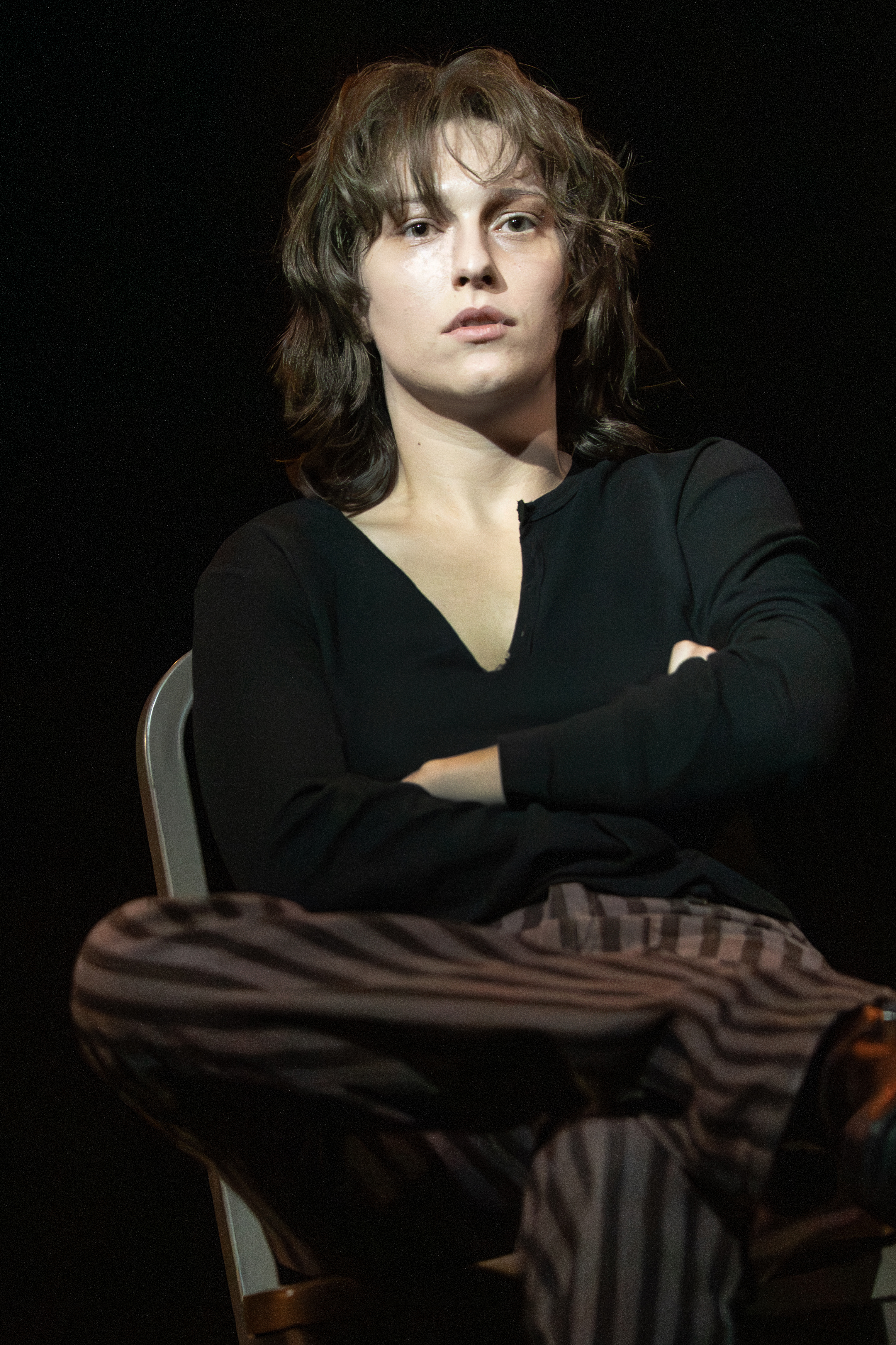 king princess as lisa wearing a black v neck long sleeved shirt and striped pants sitting on stage in girl interrupted