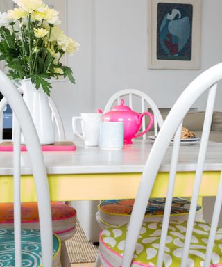 A close-up shot of a white dining table with white mugs, a white vase with yellow flowers, and a pink tea pot on it, with a yellow edge and white chairs with blue and green cushions on it