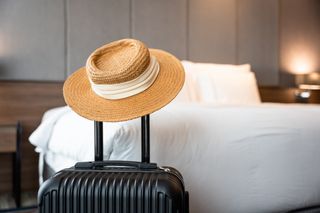 a suitcase with a straw hat on the handle, situated in a hotel room with a white bed in the background