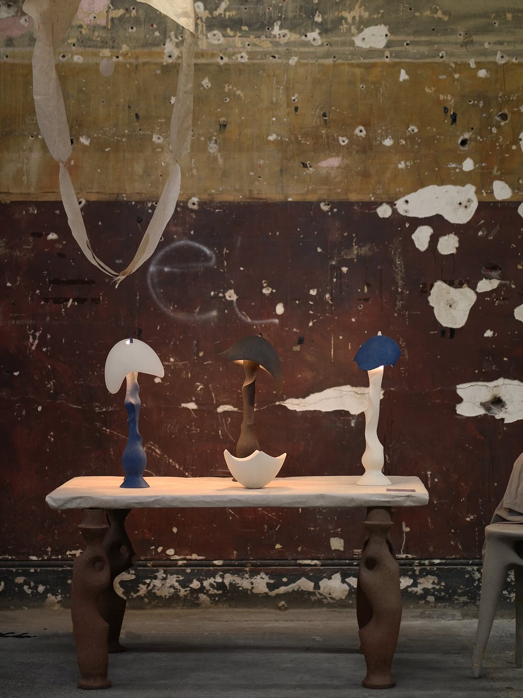 On the backdrop of a peeling brown wall, three lamps and a cup rest on a table with wobbly legs
