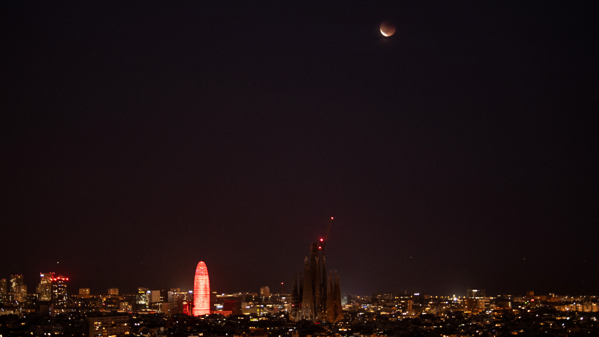 An almost fully eclipsed moon is pictured hanging in the dark sky over a city skyline. Tall buildings are visible near the center of the shot. The moon's lower crescent is brightly lit with the rest bathed in deeper shadow.
