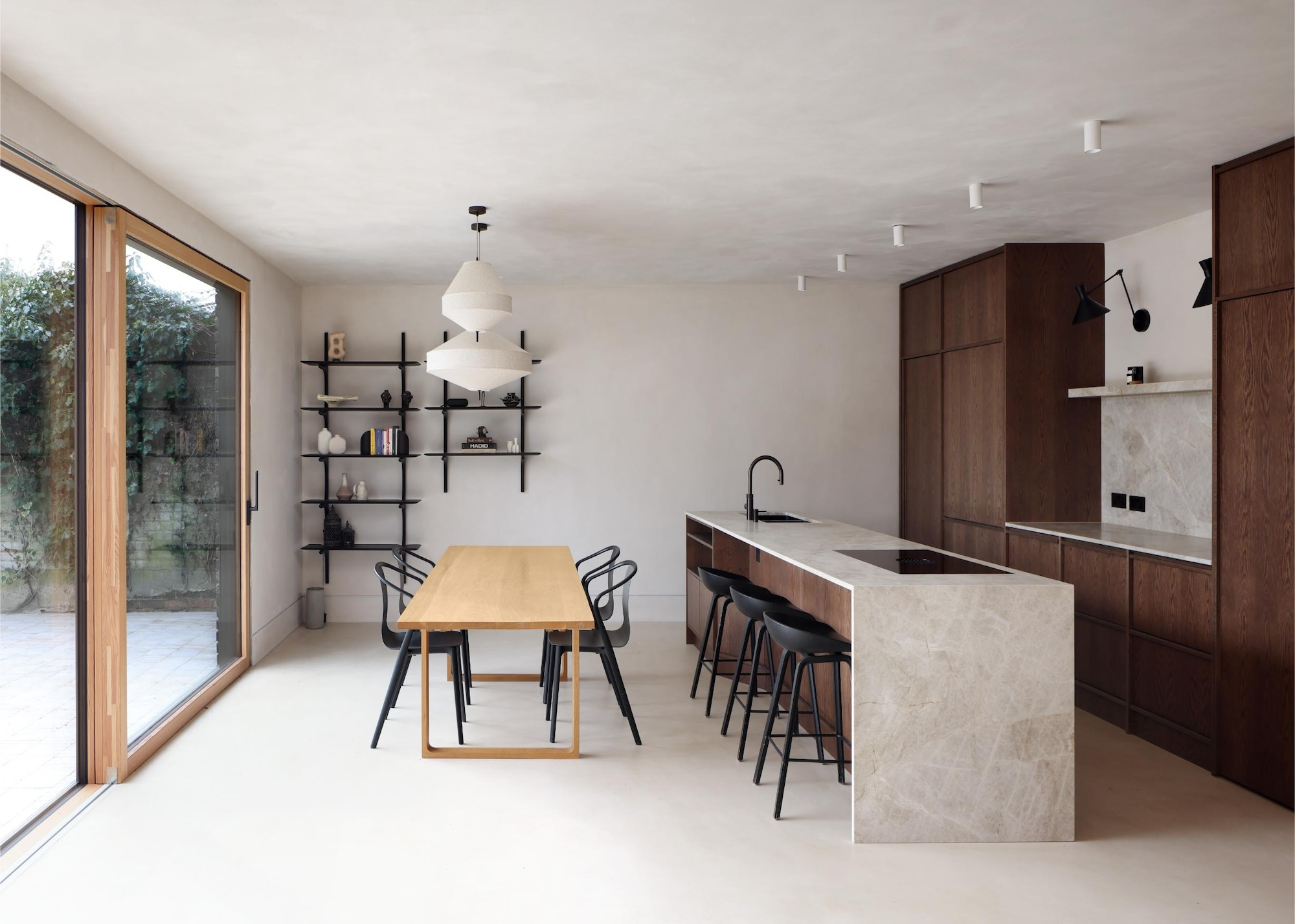 A neutral kitchen with wood-framed sliding patio doors with a light wood dining table and dark woof kitchen cabinets with a kitchen island that has black stools running along the side that is next to the dining area