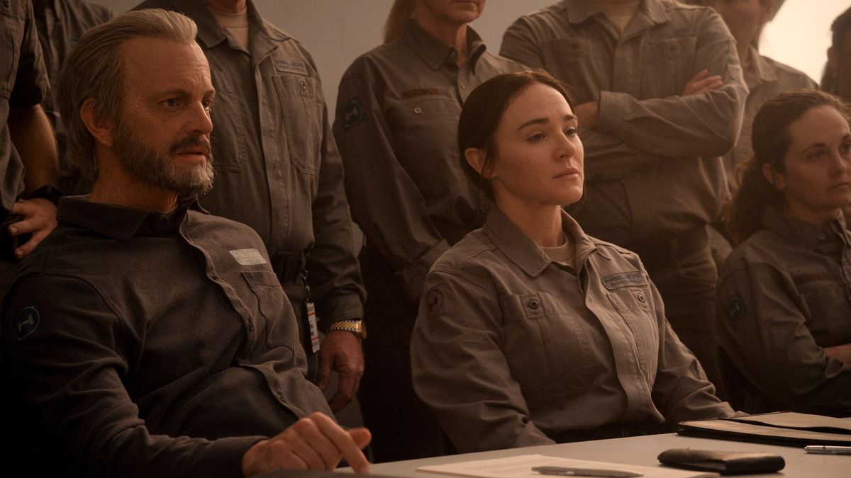 Sitting at a table are three people with very serious expressions. In the background there are more people standing, some with their arms crossed. They are all wearing the same gray jumpsuit.