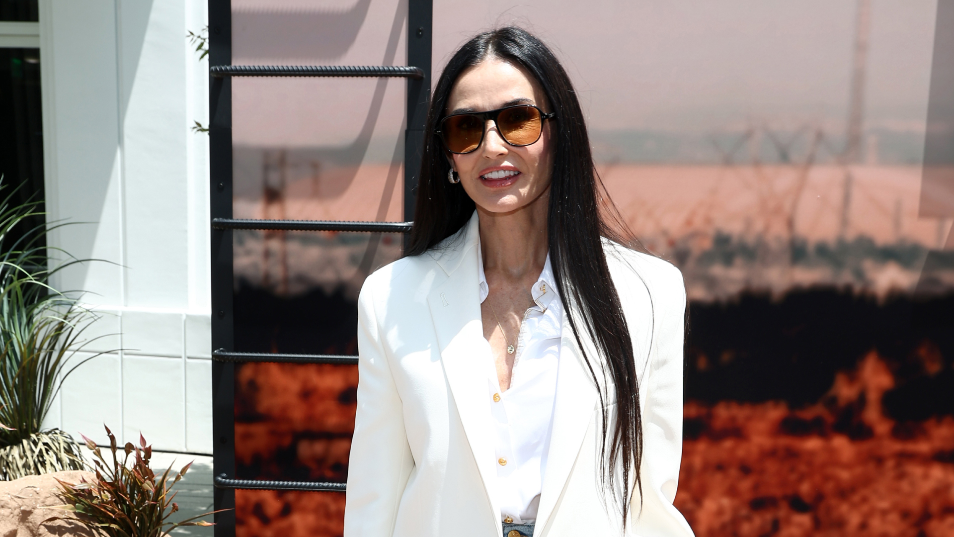 Woman with long straight hair wearing sunglasses, a white blazer and a white button up shirt.