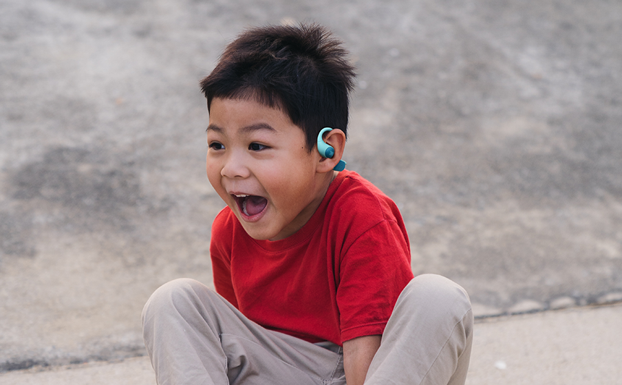 A young boy with black hair and a red t-shirt yelling happily while wearing JBL Junior Free headphones