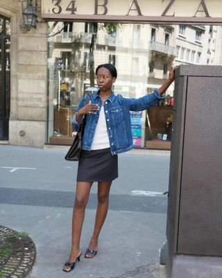 Woman standing on sidewalk wearing white t-shirt, denim jacket, black miniskirt and black heeled sandals.