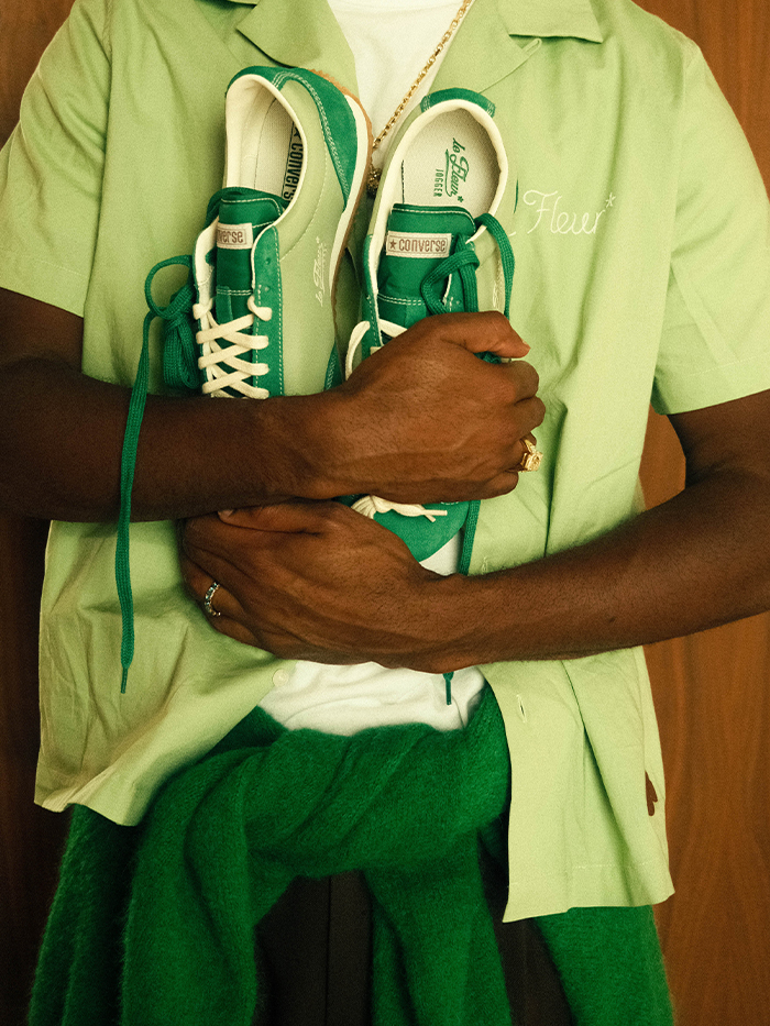 Tyler the Creator hold a green pair of the Converse Jogger shoes while wearing a green button-down and white t-shirt with a green sweater tied around his waist.