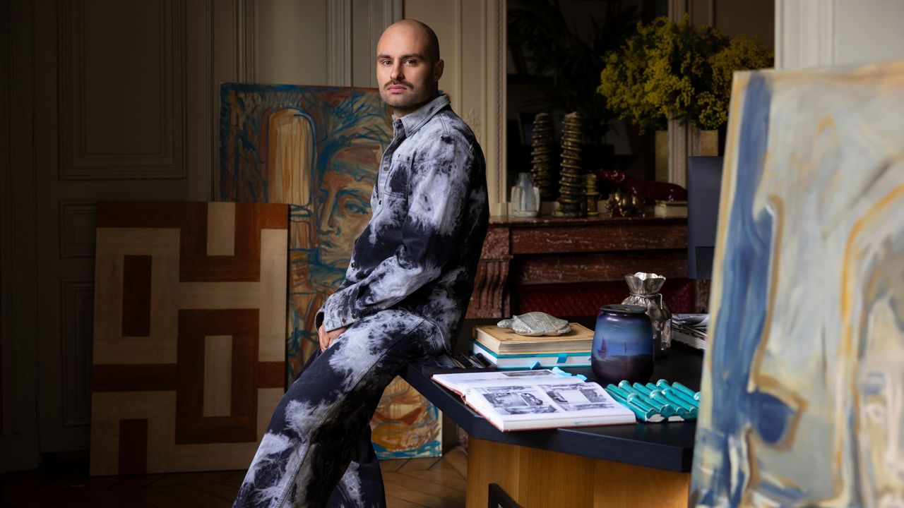 A young man dressed in an abstractly decorated 2-piece outfit in the tones of blue and light blue sits in a studio filled with pastel-shaded canvases on a wooden table covered in objects.