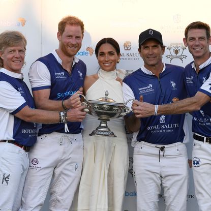 Prince Harry and Meghan Markle posing with polo players holding a trophy in front of a white background