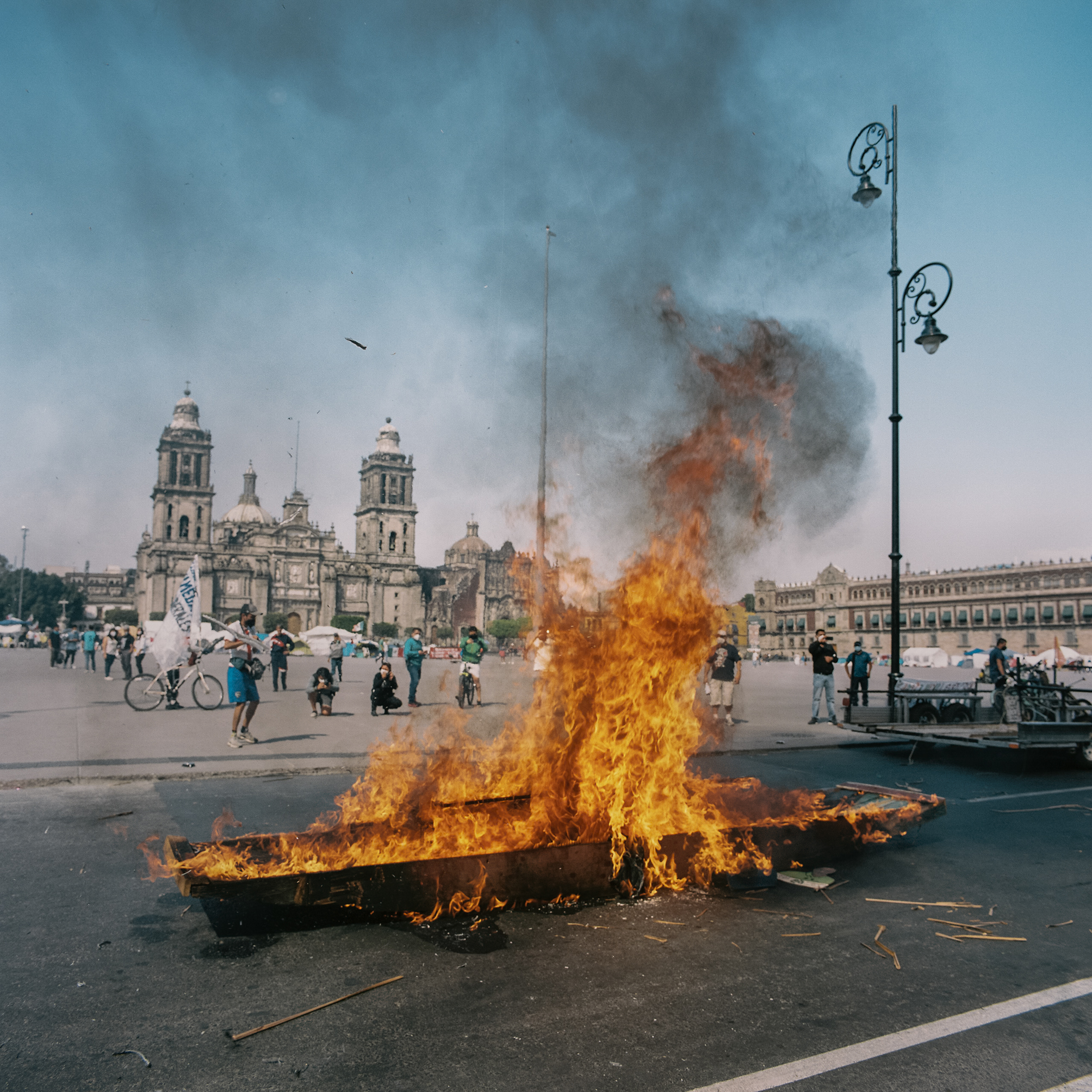 A wooden boat engulfed in bright flames burns in a large public plaza in Mexico City, with smoke rising into the sky while bystanders observe and historic buildings stand in the background.