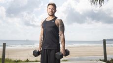 Luke Zocchi on a beach holding two dumbbells