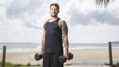 Luke Zocchi on a beach holding two dumbbells