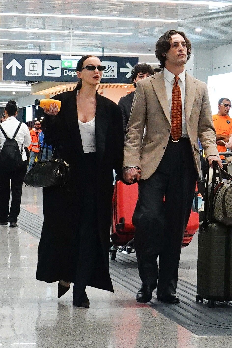 Damiano David wears a tie and blazer as he walks through the airport with Dove Cameron.