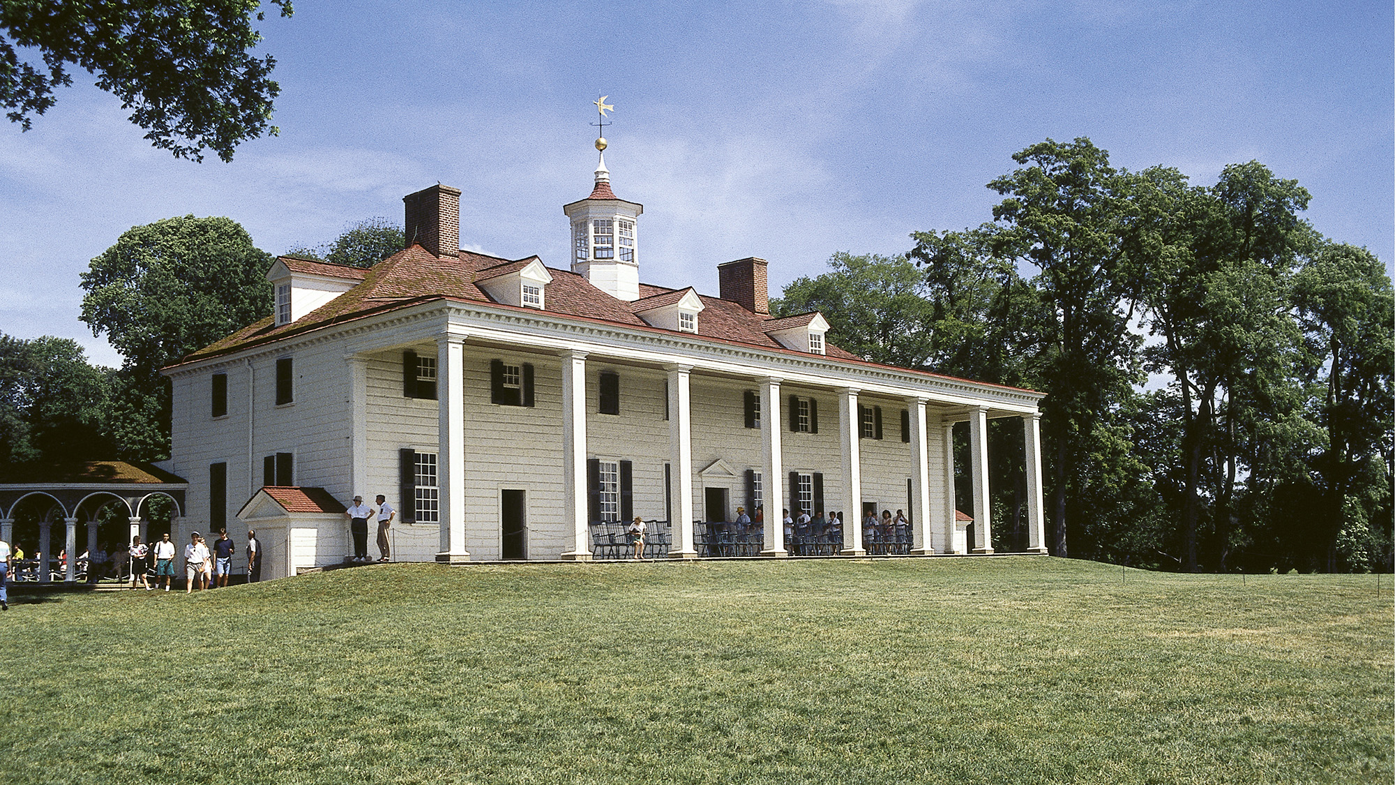 A white structure with pillars sits on a grassy hill.