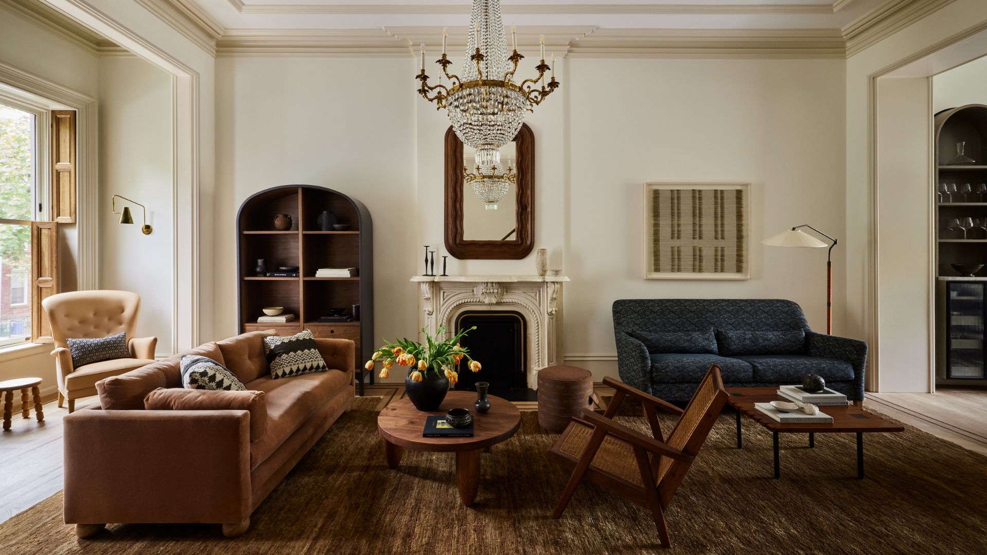 A beautiful modern living room with a wooden rattan chair, a blue sofa, a dusky peach couch, a yellow armchair, a round coffee table, a glass chandelier, a stone mantel over a fireplace