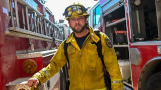 Max Thieriot as Bode standing between two fire trucks in fire gear on Fire Country.