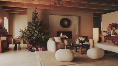 Warm, minimalist holiday living room featuring a wild evergeen dressed in red ornaments, a curved boucle armchair and ottomans, mid-century-style wooden coffee table, and a travertine fireplace