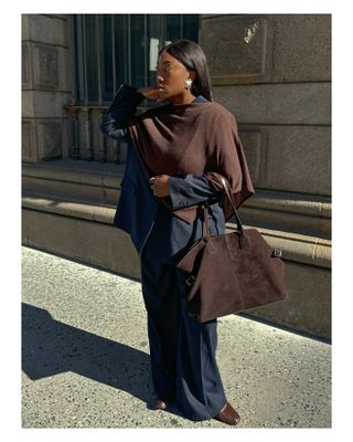 A woman wears a chocolate brown cape, navy pinstripe pants suit, brown shoes, and a brown suede tote bag.