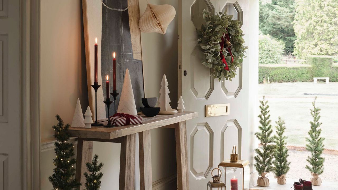 An entryway decorated for Christmas with ceramic trees on a side board, and a wreath hanging on the open door