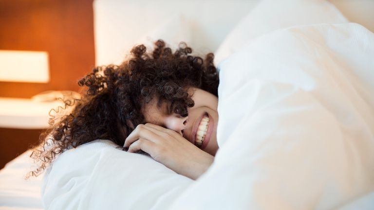 A smiling woman with dark curly hair in bed under the covers, head on a white pillow- stock photo