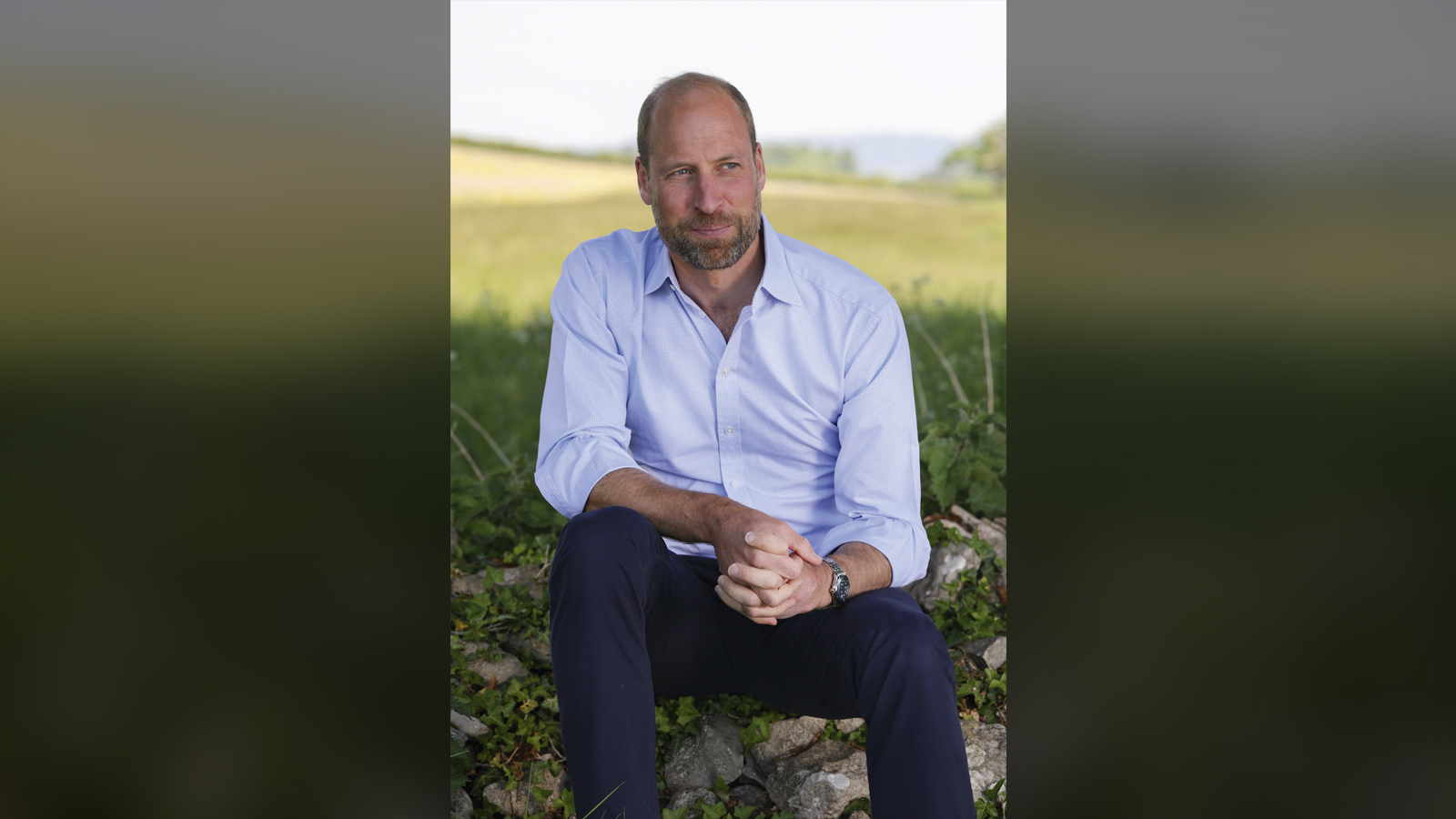 An image of HRH William, the Prince of wales, sitting on a stone wall in the countryside looking wistful