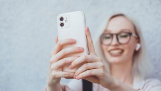 Blonde woman wearing glasses holding up phone for selfie whilst wearing earbuds