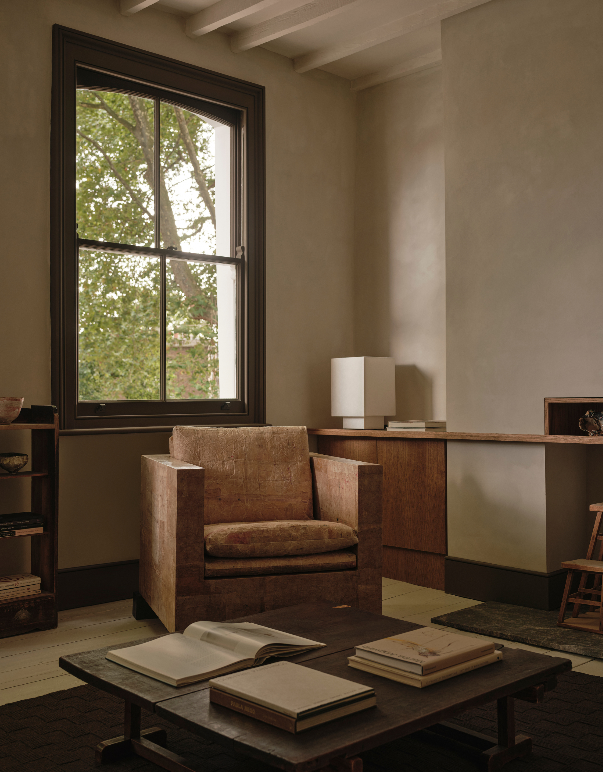 A living room with a bright window, a textured armchair, a coffee table with books, a square white table lamp, and ceramics