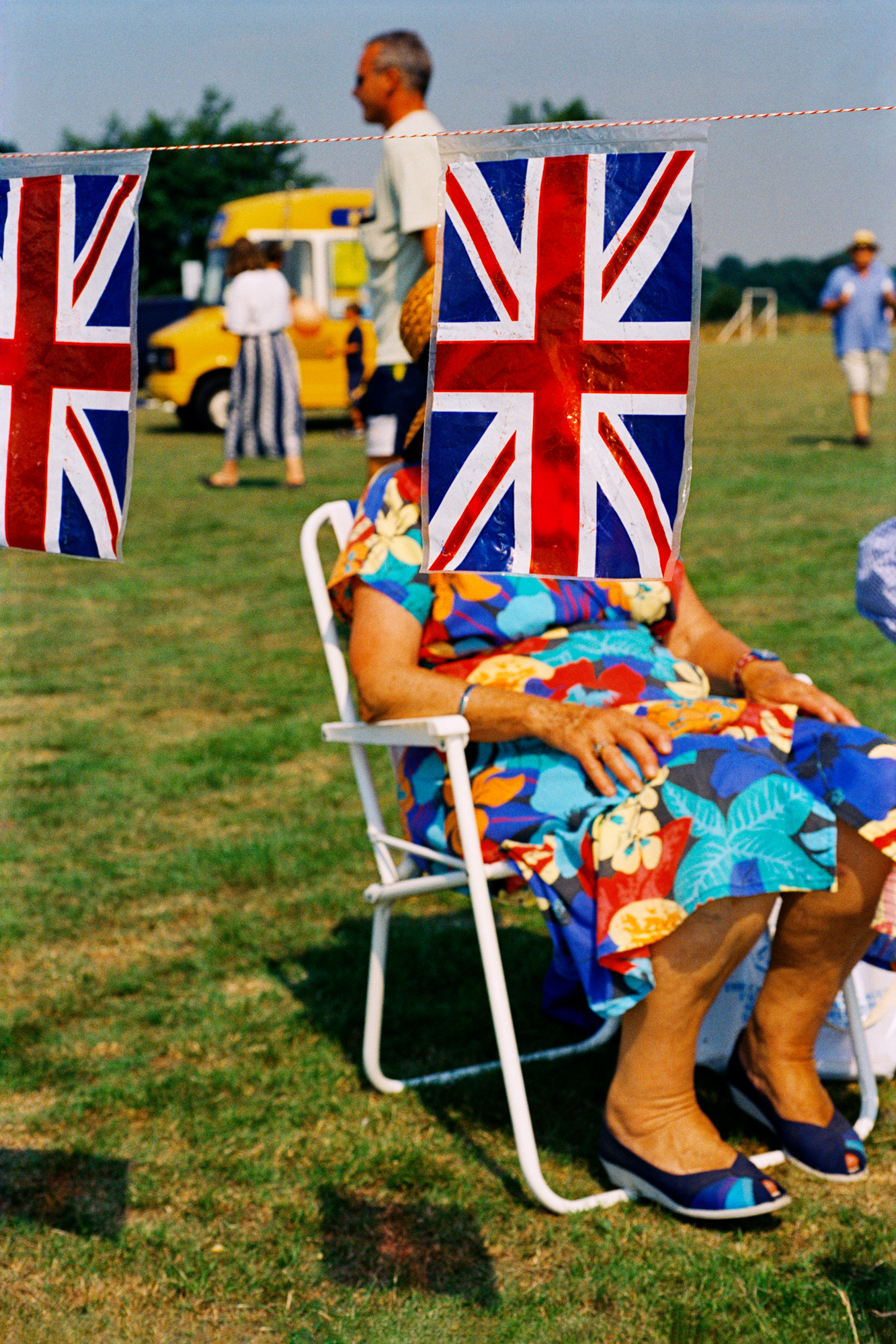 A woman in a brightly patterned dress sits in a folding chair on a grassy field, partially obscured by two hanging plastic Union Jack flags, with other people and a yellow van visible in the background.