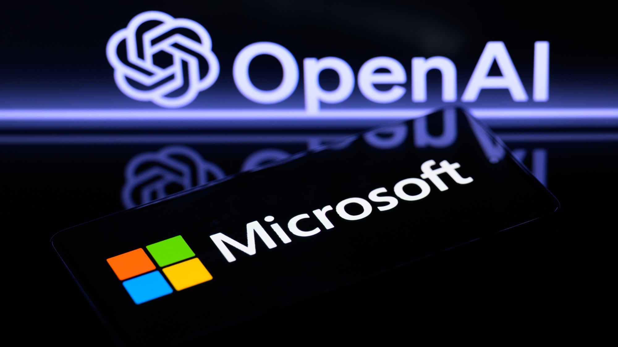 The Microsoft logo is being displayed on a smart phone, with the OpenAI logo visible on the screen in the background.