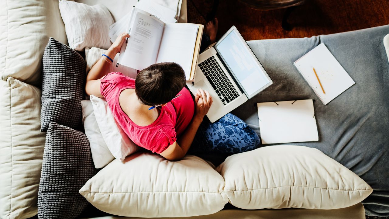 Aerial view of a woman sitting on her sofa and working on her laptop while surrounded by paperwork.