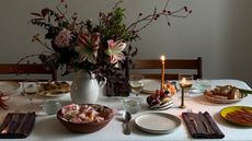 Image of a table with a white tablecloth set with plates, cutlery, dark napkins, and plates of food. There is a large floral arrangement in a white vase and candles in the center of the table.