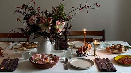Image of a table with a white tablecloth set with plates, cutlery, dark napkins, and plates of food. There is a large floral arrangement in a white vase and candles in the center of the table.