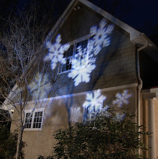 Electric LED Projector Light casting white snowflake shapes onto a home facade