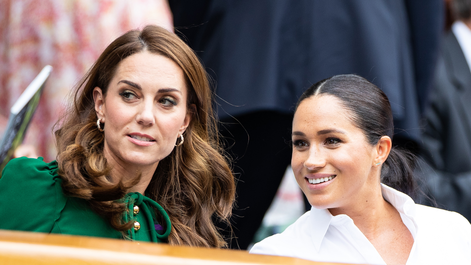 Princess Kate Middleton wears a green dress and has long wavy brown hair and Duchess Meghan Markle wears a white shirt and wears her hair in a ponytail