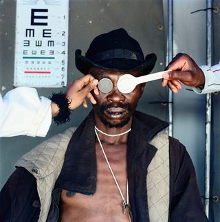Ex-prisoner Joseph having his eyes tested. He is shirtless and wearing a black and beige jacket, a black hat and a long necklace