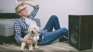 A woman in jeans sits on the floor with a small white dog next to a speaker