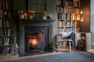 A lounge with a lit fireplace with a grey cushion and a painted black surround, two bookcases and a wooden chair with a grey cushion and grey throw draped over it