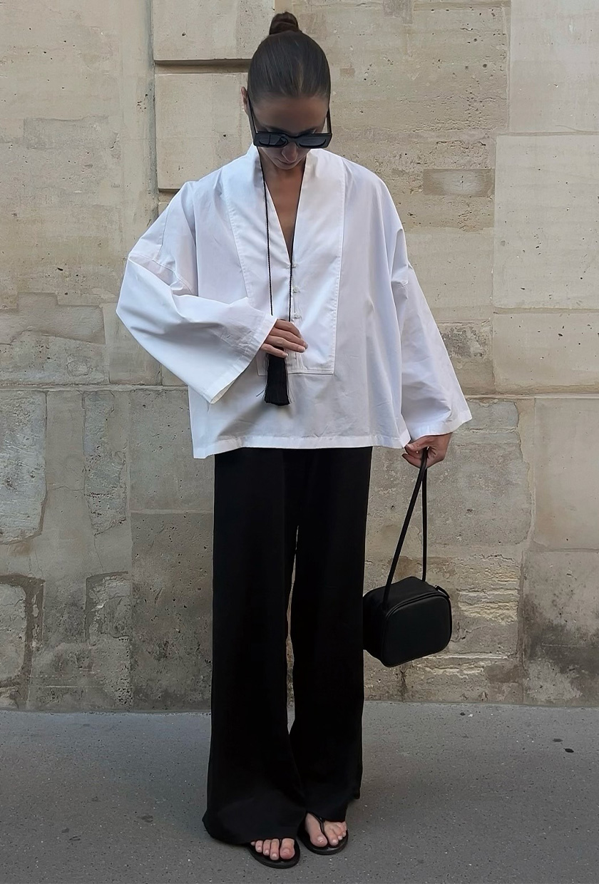 A woman in Paris wearing a white caftan blouse styled with black wide-leg trousers, black flip-flops, a black tassel necklace, and a black bag.