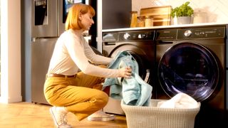 Redhead woman placing laundry from basket into dark washing machine in contemporary kitchen with wooden floor