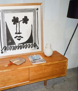 View of framed artwork by Arkitaip and Hôtel Magique. The black and white piece features a face with flowers for eyes and sits on top of a wooden sideboard with other items on top in a space with white walls and grey flooring