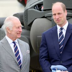 King Charles wears a gray suit and smiles at son Prince William, who has a bald head and is wearing a navy suit