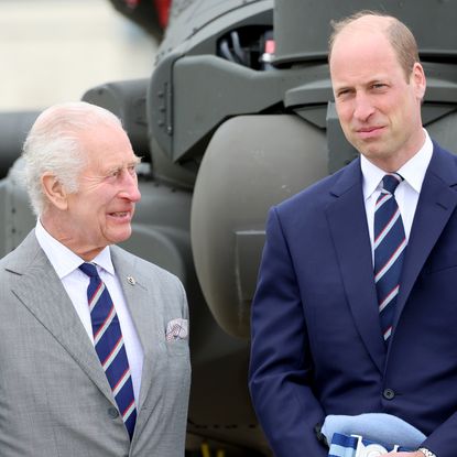 King Charles wears a gray suit and smiles at son Prince William, who has a bald head and is wearing a navy suit