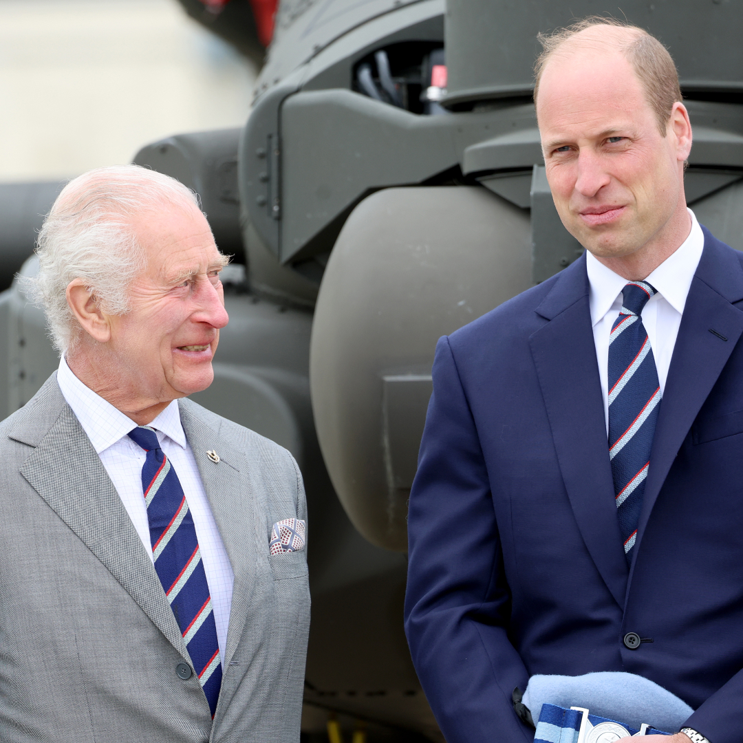 King Charles wears a gray suit and smiles at son Prince William, who has a bald head and is wearing a navy suit
