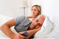 A couple, perhaps in their 50s, rests in bed. The man is looking at his cell phone and turned away from his wife or partner.