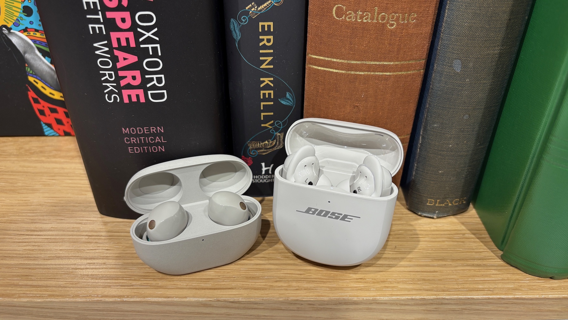The Bose QuietComfort Ultra Earbuds (2nd Gen) and Sony WF-1000XM5 earbuds in their charging cases on a shelf in front of some books.